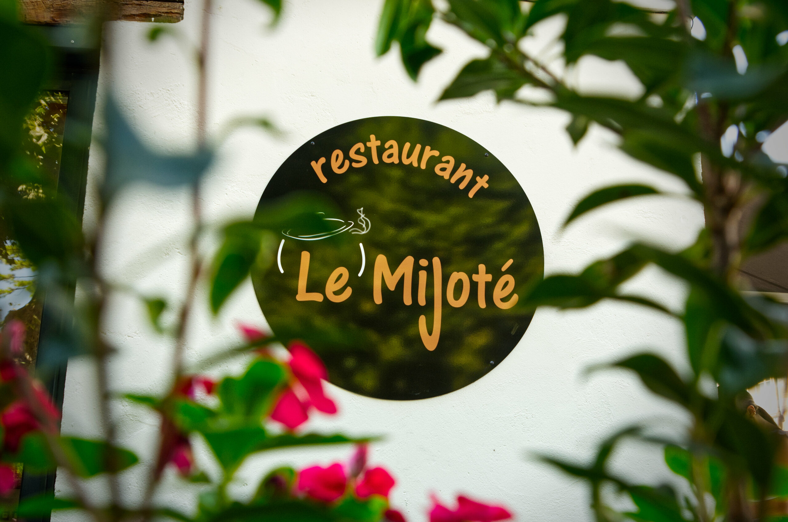 Enseigne du restaurant Le Mijoté sur la terrasse, vu au travers des fleurs.